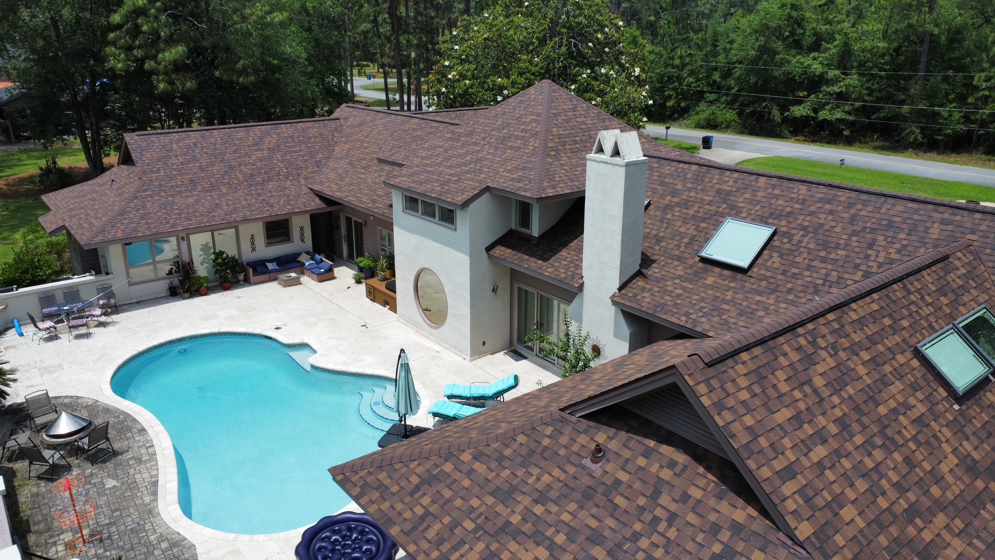 Aerial view of a large house with a brown shingle roof, a swimming pool, outdoor seating, and surrounding trees.