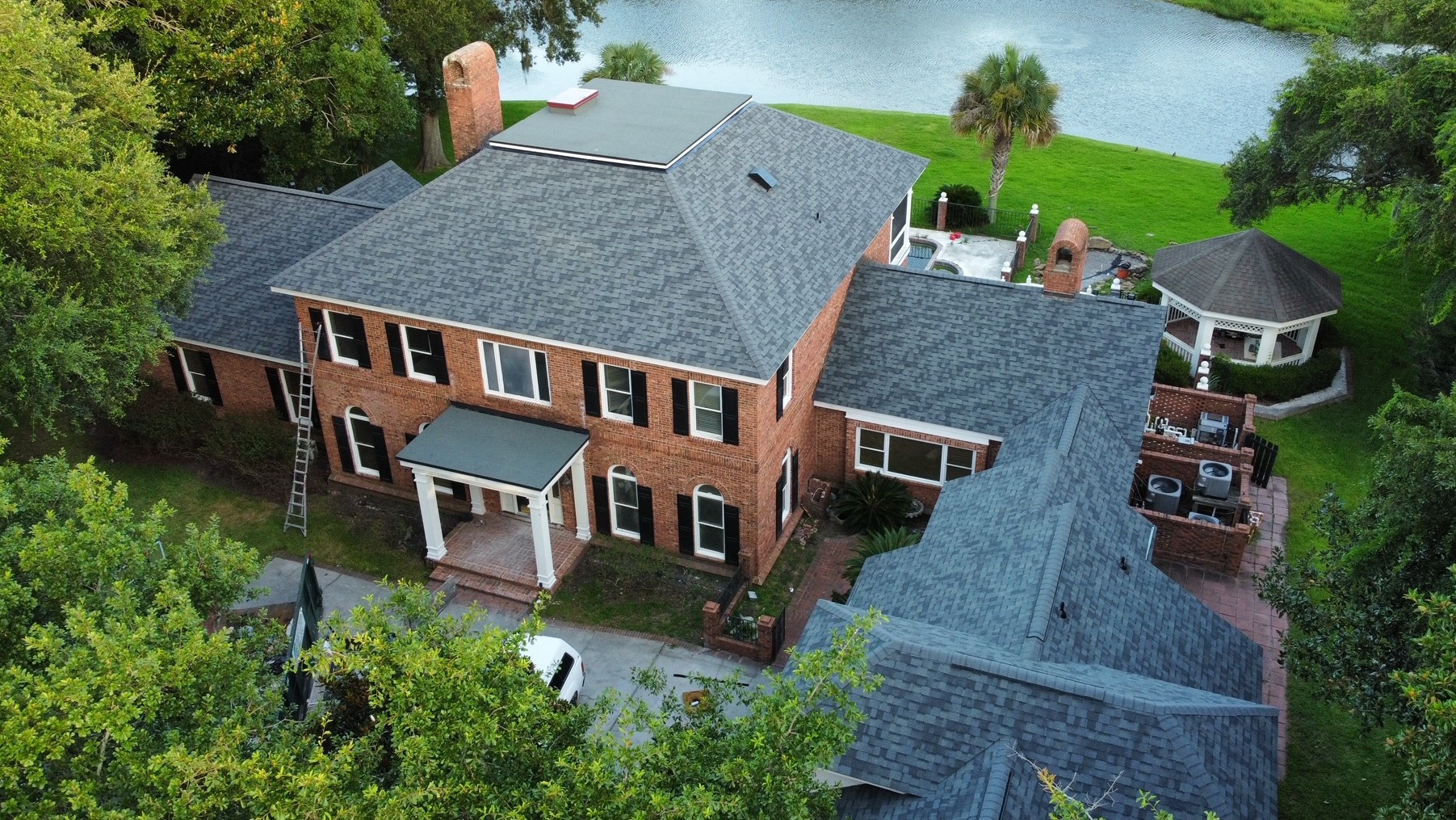 Aerial view of a large two-story brick house with gray shingle roof, surrounded by trees, a gazebo, and a lake in the background.