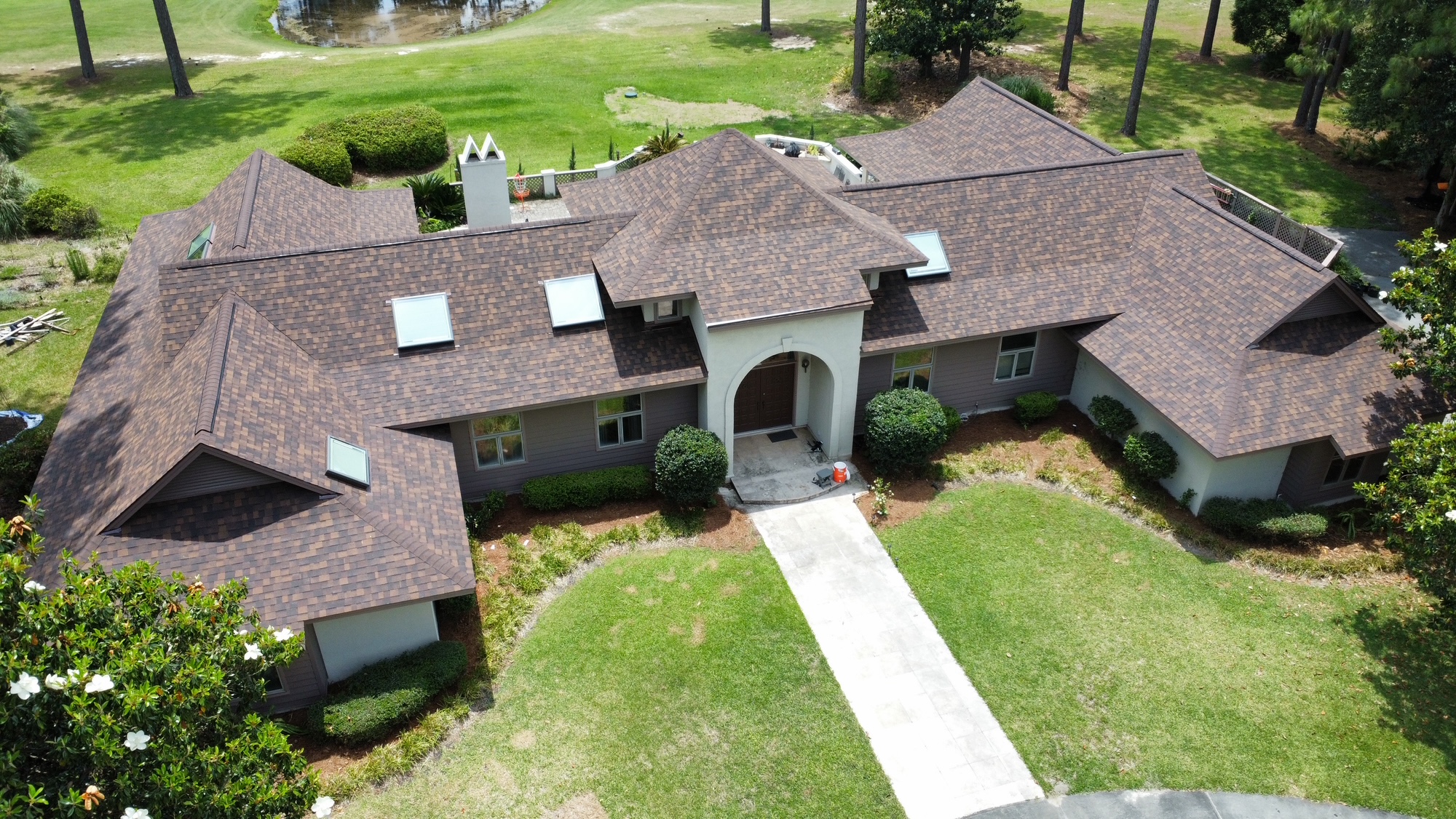 Aerial view of a large, single-story house with a brown shingle roof, surrounded by green lawns, shrubs, and trees.