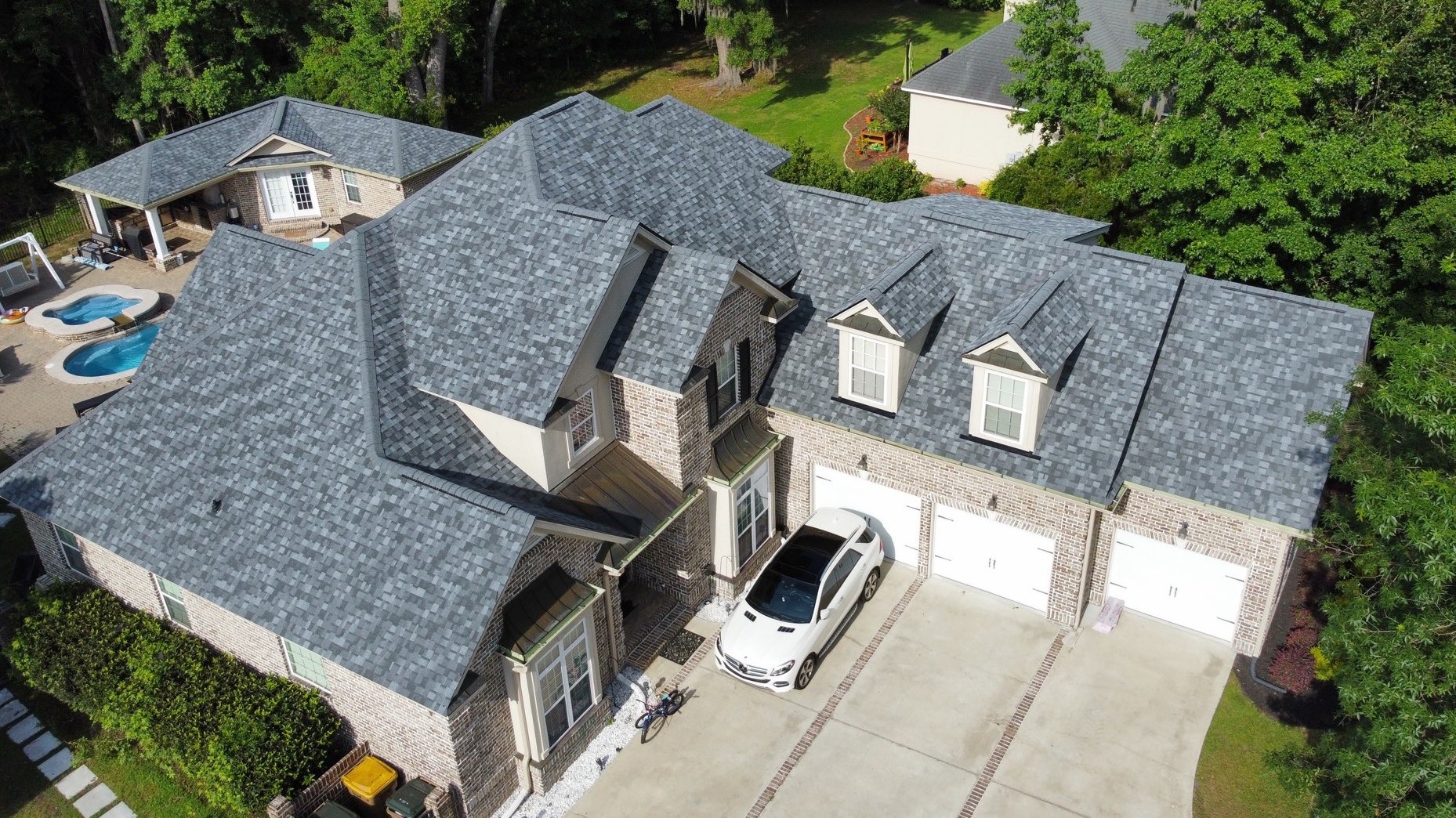 Aerial view of a large brick house with a gray shingle roof, a white car parked in the driveway, and a backyard with a pool and greenery.