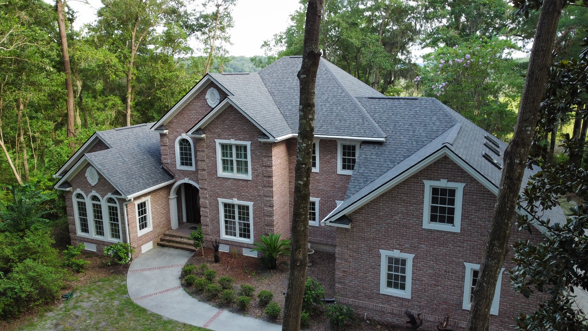 Two-story brick house with gray shingle roof, multiple windows, and a curved driveway, surrounded by trees and greenery.