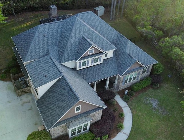Aerial view of a two-story suburban house with a grey shingled roof, brick and siding exterior, driveway, front porch, and surrounding lawn and trees.
