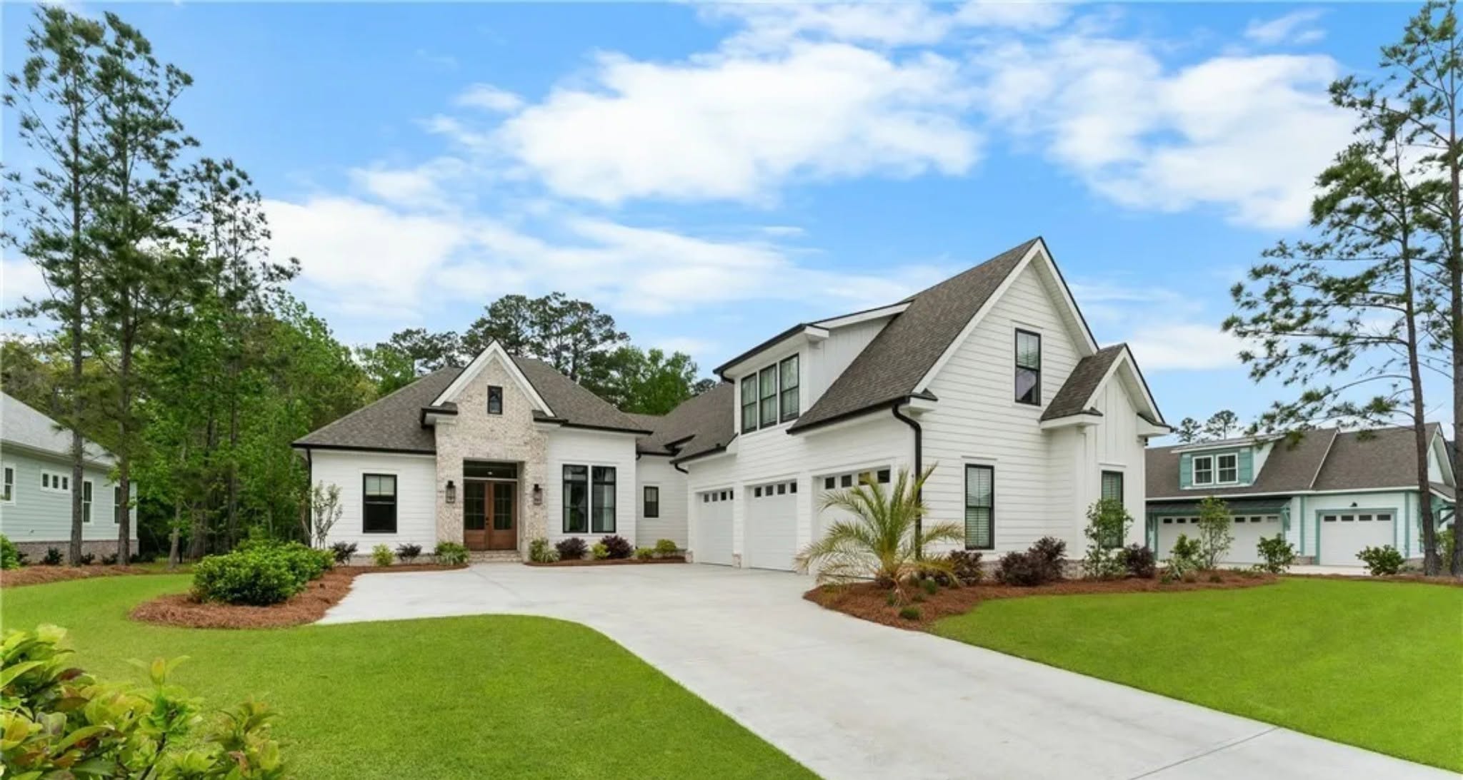 Modern two-story white house with attached three-car garage, gabled roof, manicured lawn, and surrounding trees under a blue sky with scattered clouds.