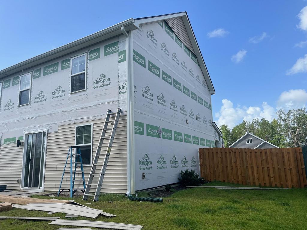 A house under renovation with siding removed on one side, revealing GreenGuard insulation wrap, ladders leaning against the wall, and construction materials on the grass.